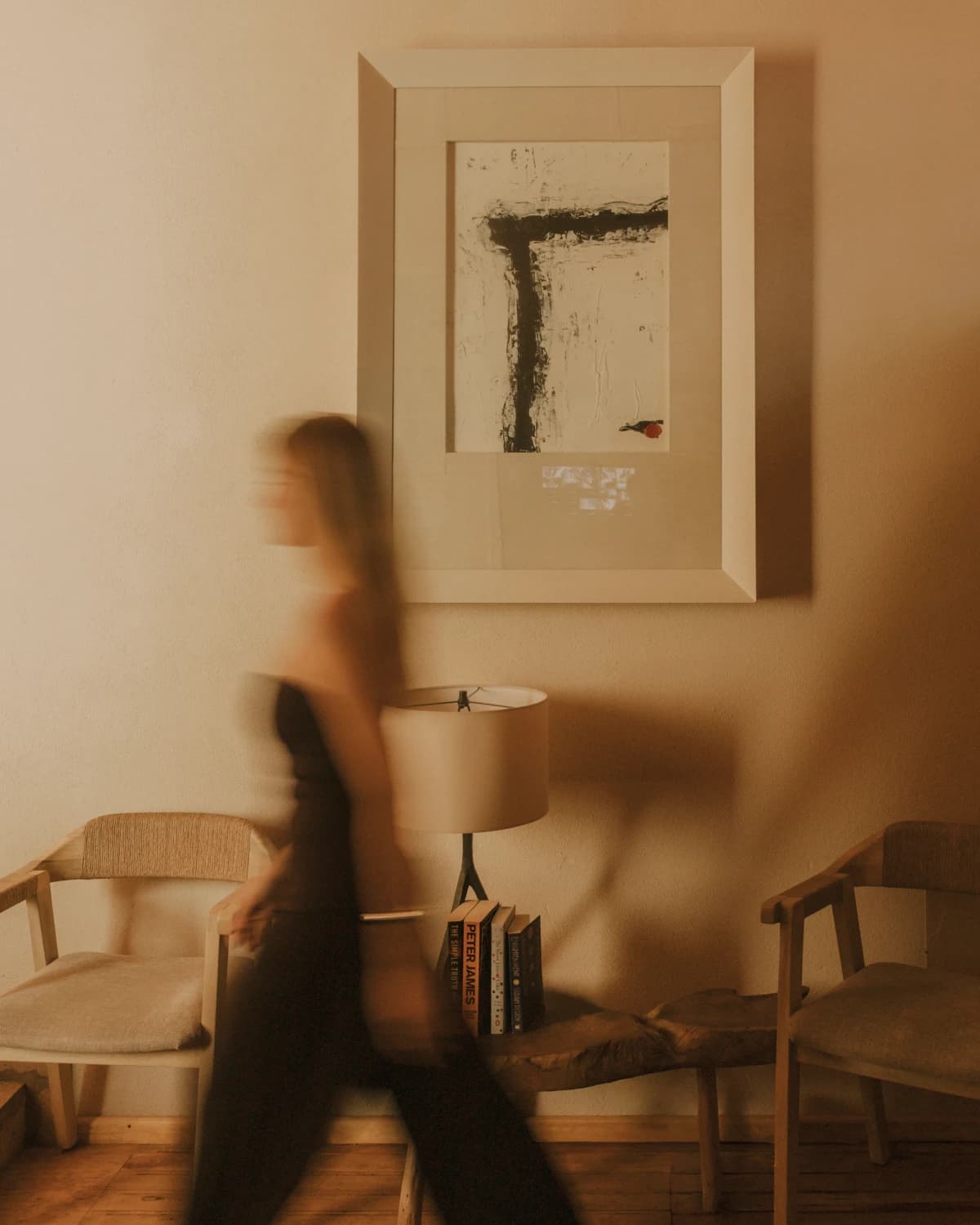 A hallway at Orchid House Coyoacán with a framed abstract painting, a bench with books beneath a lamp, and two wood-and-rope armchairs, as a guest walks past